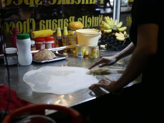 In una bancarella di cibo notturna, una persona stende una sottile sfoglia di pasta circolare su un bancone di metallo, circondata da ingredienti come banane e mais.