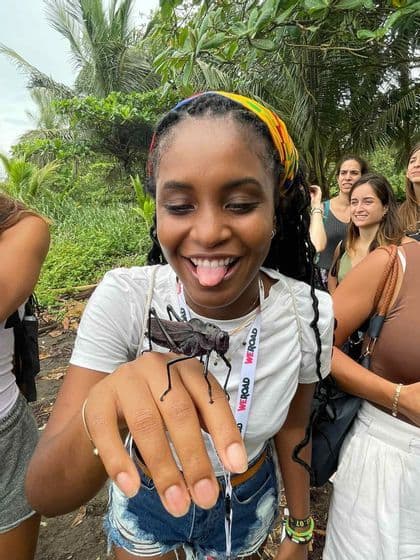 Una donna sorridente durante un viaggio di gruppo WeRoad tiene giocosamente un grande insetto nero sulla mano, all'aperto in un ambiente lussureggiante.