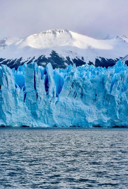 A massive blue glacier rises from the dark water, with snow-covered mountains in the background.