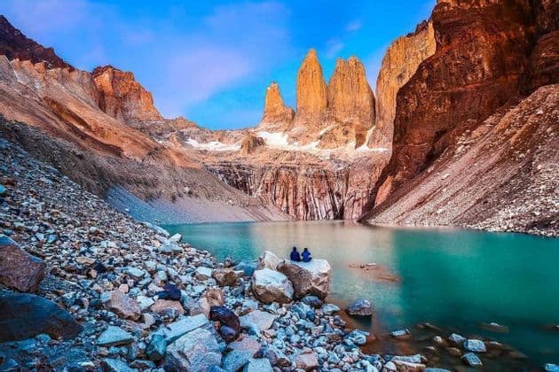 Two people sit on a large boulder on the shore of a turquoise lake at the base of dramatic, sunlit mountain spires.