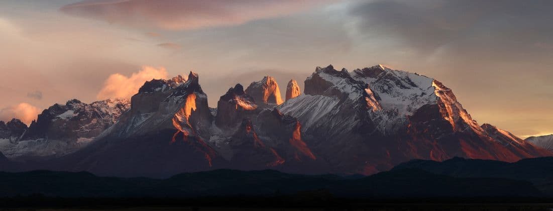 Ein Panoramablick auf eine schneebedeckte Bergkette mit zerklüfteten Gipfeln, beleuchtet vom warmen Licht des Sonnenaufgangs oder Sonnenuntergangs.