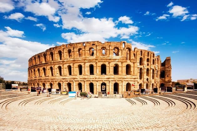 Eine Weitaufnahme eines großen, antiken Steinamphitheaters mit gewölbten Öffnungen unter blauem Himmel mit weißen Wolken.
