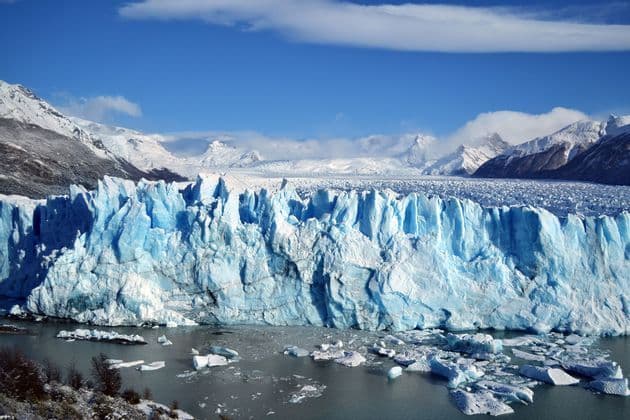 Une imposante paroi glaciaire bleue rencontre un plan d'eau où flottent des blocs de glace, avec des montagnes enneigées en arrière-plan.