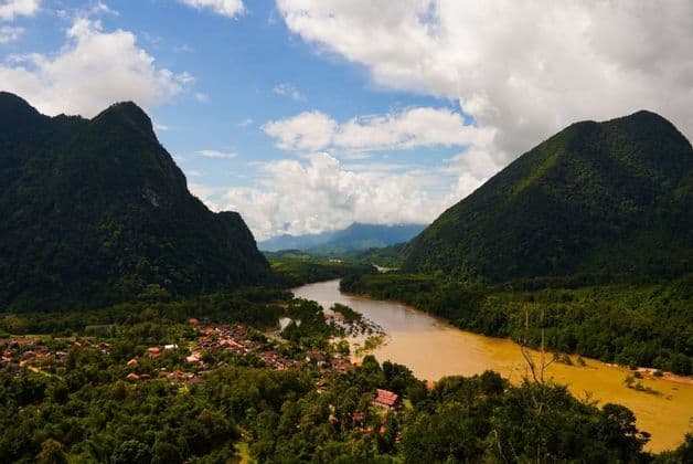 Vue aérienne d'une rivière boueuse traversant une vallée entre deux grandes montagnes couvertes de forêts, avec un petit village visible sur la rive.