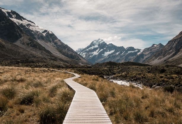 A winding wooden boardwalk leads through a grassy valley towards tall, snow-capped mountains under a cloudy sky.
