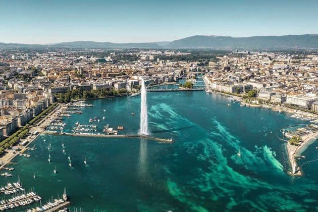 An aerial view of a large water fountain spraying from a turquoise lake, with a cityscape and boat-filled harbor along the shore.