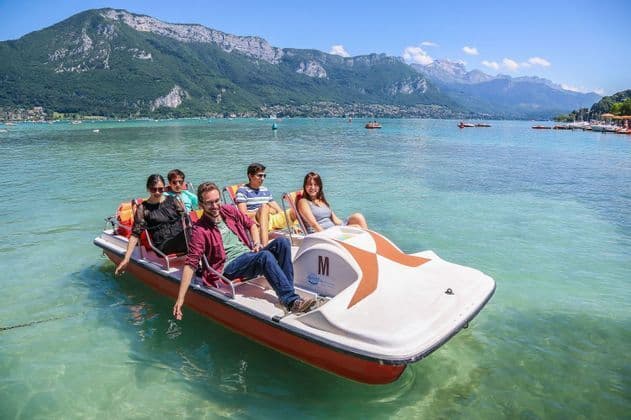 A WeRoad group trip of five people relaxing on a pedal boat on a clear lake with large, green mountains in the background.