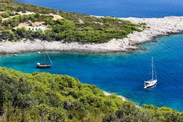 Zwei Segelboote ankern in einer türkisfarbenen Bucht neben einer felsigen Küste mit grünen Bäumen, von oben gesehen.