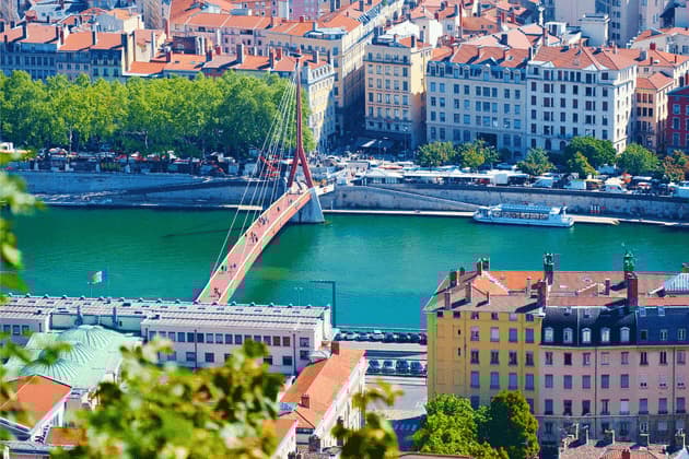 Vue plongeante d'un pont piétonnier rouge traversant une rivière verte dans une ville européenne avec des bâtiments aux toits de tuiles en terre cuite.
