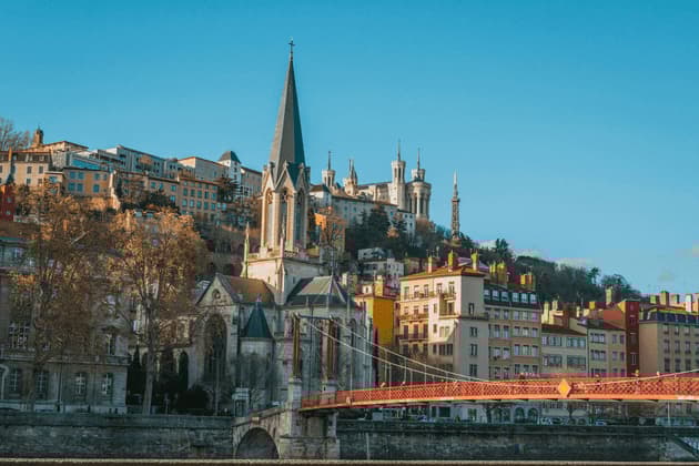 Une ville européenne historique construite sur une colline, avec une grande église et un pont rouge au premier plan sous un ciel bleu clair.