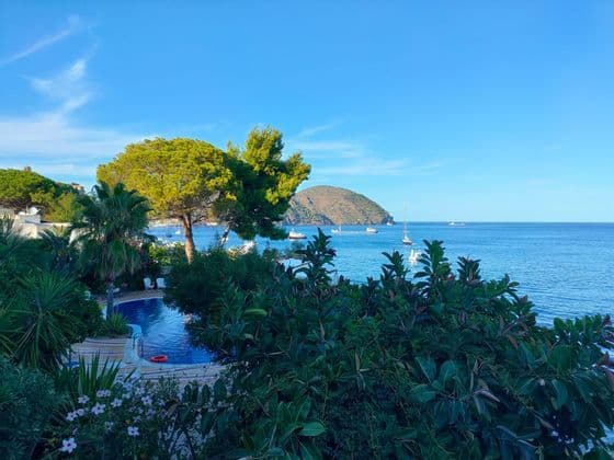 Una piscina immersa nel verde lussureggiante si affaccia su un mare blu con barche e un'isola lontana sotto un cielo sereno.