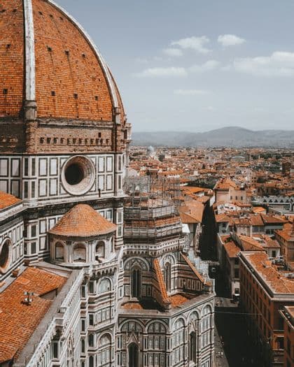 Veduta dall'alto di una grande cupola di cattedrale con tegole rosse che si affaccia su un paesaggio urbano con tetti simili e montagne lontane, sotto un cielo azzurro.