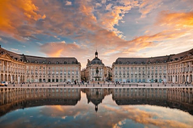 Un grand bâtiment historique se reflète parfaitement dans un miroir d'eau sur une place de ville sous un ciel de coucher de soleil coloré.