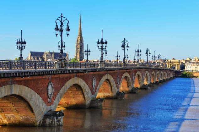 Un pont de briques voûté avec des lampadaires ornés enjambe une large rivière face à un paysage urbain où se dresse un haut clocher d'église.