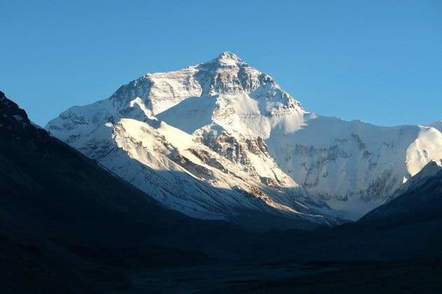 Una cima montuosa alta e innevata è illuminata dal sole contro un cielo blu limpido, sorgendo da una valle in ombra.