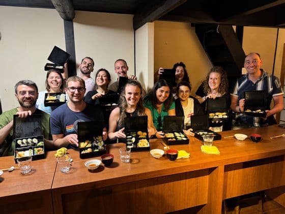 Un gruppo WeRoad di persone sorridenti riunite attorno a un bancone di legno, che mostrano bento box aperte piene di sushi.