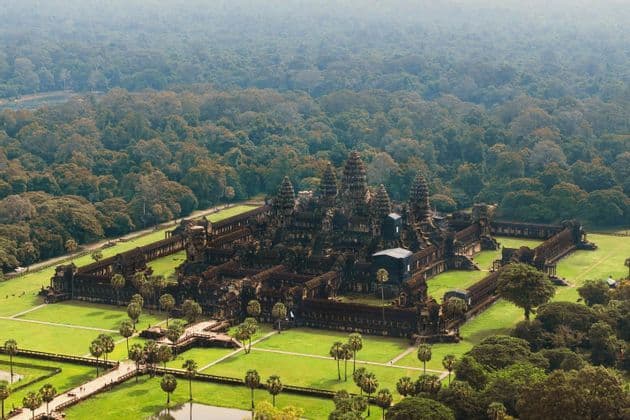 Una vista aerea di un grande e antico complesso di templi in pietra con torri, circondato da prati verdi, palme e una vasta giungla.