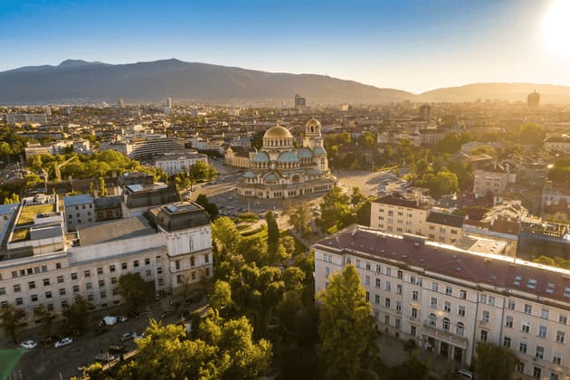 Vista aérea de una extensa ciudad, con una gran catedral de cúpulas doradas en el centro y montañas de fondo al atardecer.
