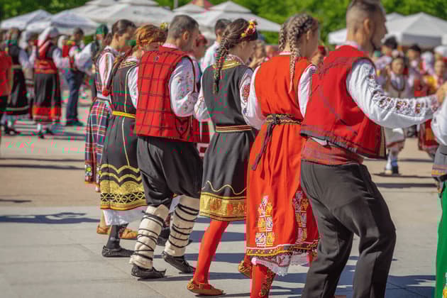Una fila de bailarines folclóricos con coloridos trajes tradicionales bordados actúan al aire libre.