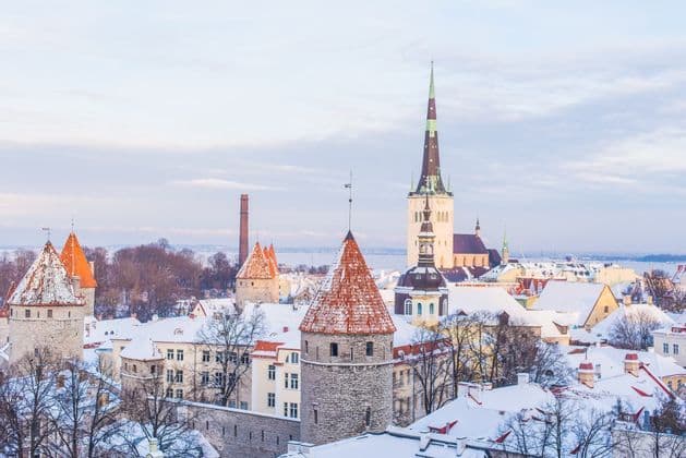 Uno skyline di una città storica in inverno, con tetti coperti di neve, torri di pietra con tetti rossi e un'alta guglia di chiesa.