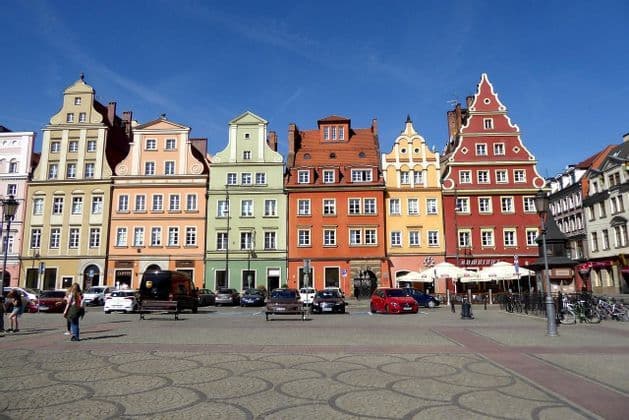 Una hilera de coloridos edificios históricos con tejados a dos aguas se alza en una plaza adoquinada bajo un cielo azul claro.