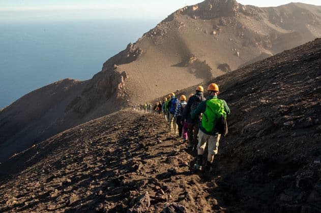 Un gruppo WeRoad con caschi e zaini fa trekking in fila su un ripido sentiero vulcanico a picco sul mare.