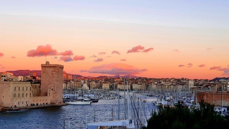 Un fort en pierre domine un port rempli de voiliers, avec une ville et des montagnes à l'arrière-plan sous un coucher de soleil coloré.