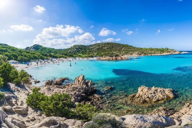 Menschen entspannen und schwimmen in einer abgelegenen Strandbucht mit türkisfarbenem Wasser, umgeben von grünen Hügeln unter einem sonnigen Himmel.