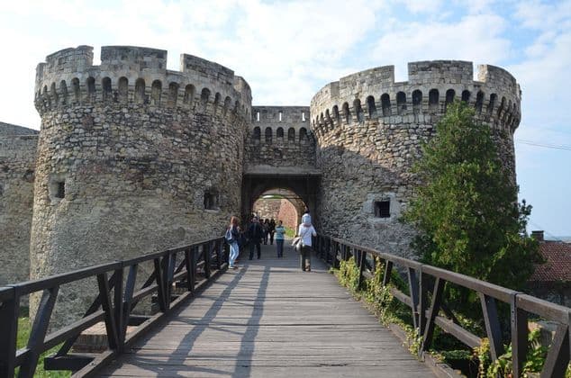 Personas caminando por un largo puente de madera que lleva a la puerta arqueada de una fortaleza medieval de piedra con dos grandes torres redondas.