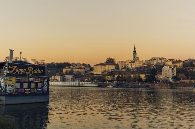Una casa flotante llamada Zappa Barka flota en un río con el horizonte de una ciudad, incluyendo un campanario de iglesia, visible al fondo al atardecer.