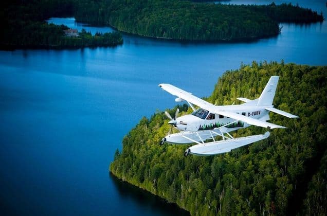 Un hydravion blanc vole bas au-dessus d'un vaste lac bleu, avec une dense forêt verte bordant les rives.