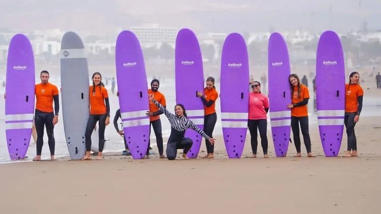 Un voyage de groupe WeRoad de personnes en combinaisons néoprène posant avec leurs planches de surf violettes sur une plage de sable au bord de l'océan.