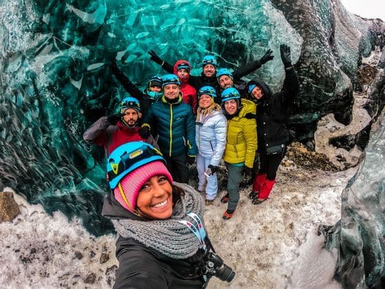 A smiling WeRoad group trip wearing helmets takes a selfie inside a vibrant turquoise ice cave.