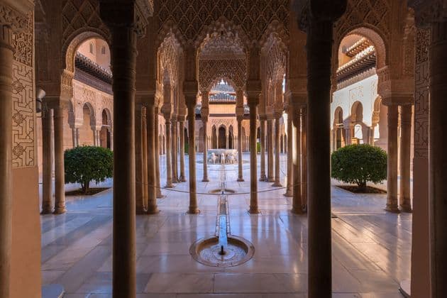 A sunlit Moorish courtyard with a central fountain, viewed from between ornately carved arches and slender columns.
