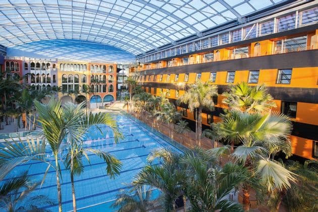 Una grande piscina coperta circondata da palme e colorati edifici dell'hotel, sotto un'enorme tetto in vetro ad arco.