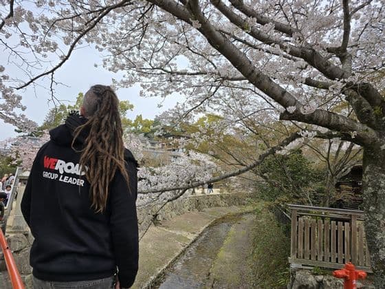 Un guía de grupo WeRoad con rastas está de pie bajo un gran cerezo en flor, observando un pequeño canal.