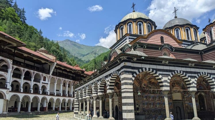 Un grand monastère aux arcades rayées et aux dômes se dresse dans une cour, au pied d'une montagne verdoyante, sous un ciel partiellement nuageux.
