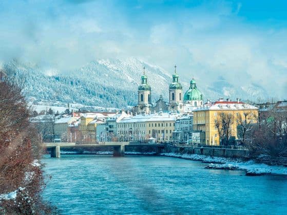Una vista di una città storica con edifici colorati e cupole di chiese verdi, lungo un fiume, con grandi montagne innevate sullo sfondo.