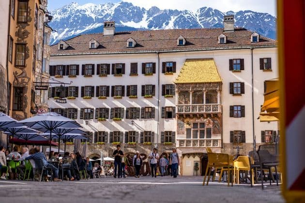 Une place de ville animée avec des gens aux terrasses de cafés, devant un bâtiment orné avec un toit doré et des montagnes enneigées.