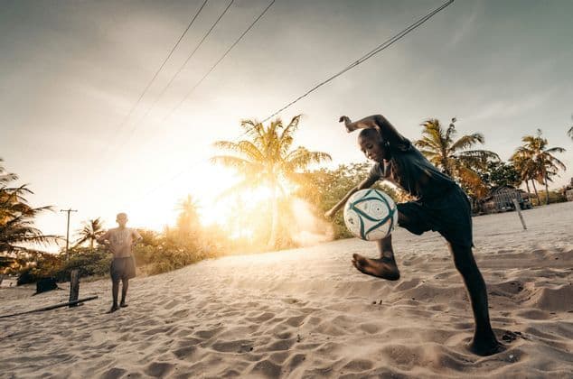 A young boy kicks a soccer ball on a sandy beach at sunset, with another boy watching in the background among palm trees.