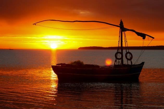 A silhouetted fishing boat floats on the water during a vibrant orange sunset, with the sun low on the horizon.