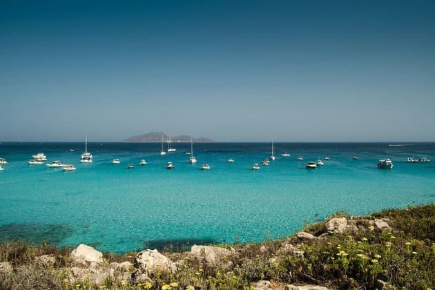 Una vista da una costa rocciosa di numerose barche ancorate in un mare calmo e turchese sotto un cielo azzurro e limpido, con un'isola in lontananza.