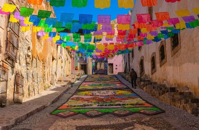 Une rue pavée décorée d'une longue tapisserie de sol colorée et de bannières de papel picado vibrantes suspendues au-dessus.