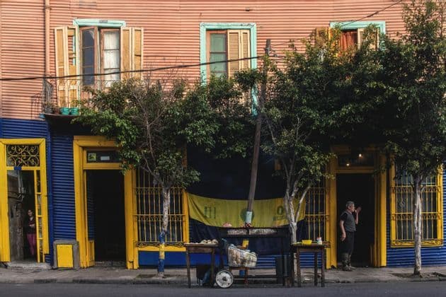 Un edificio colorato con rivestimento in lamiera ondulata rosa e blu, portoni gialli e alberi lungo il marciapiede.