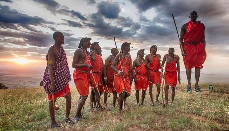 Eine Gruppe Massai-Männer in traditionellen roten Gewändern auf einem grasbewachsenen Hügel bei Sonnenuntergang, wobei ein Mann einen Hochsprung ausführt.
