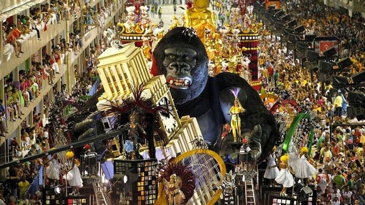 Ein riesiger Festwagen im Gorilladesign, der ein Modellgebäude trägt, bewegt sich während eines Karnevals durch eine von Zuschauern gesäumte Straße.