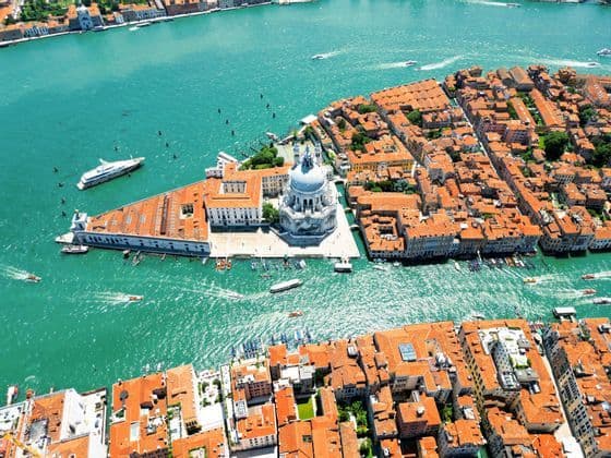 Vista aerea di una città con tetti di tegole arancioni e un grande edificio a cupola bianco circondato da acqua turchese e barche.