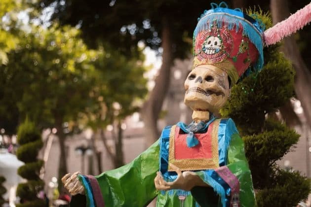 Un squelette décoratif vêtu d'un costume coloré et d'une coiffe haute et élaborée ornée d'un emblème de crâne, se dresse dans un parc.