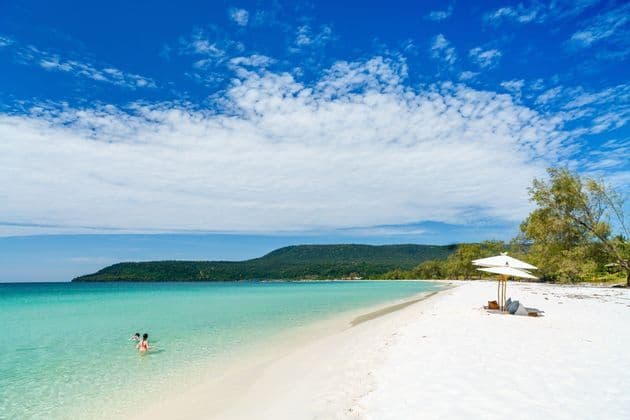 Una vasta distesa di spiaggia di sabbia bianca incontra acque limpide e turchesi, con una grande collina verde in lontananza sotto un cielo azzurro.