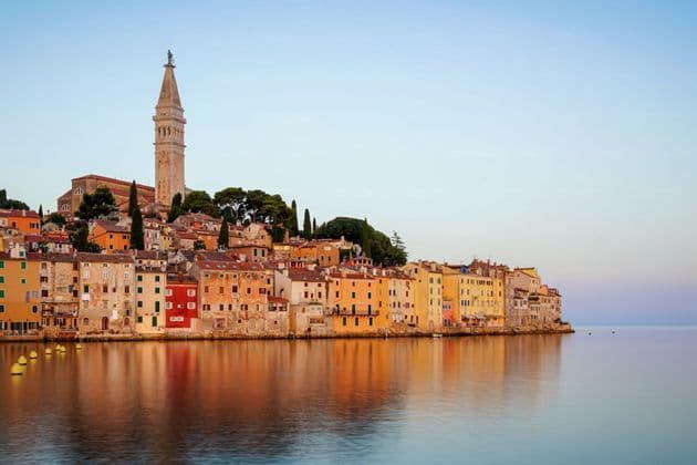 A coastal town with colorful buildings and a prominent bell tower reflecting in the calm water under a clear sky.